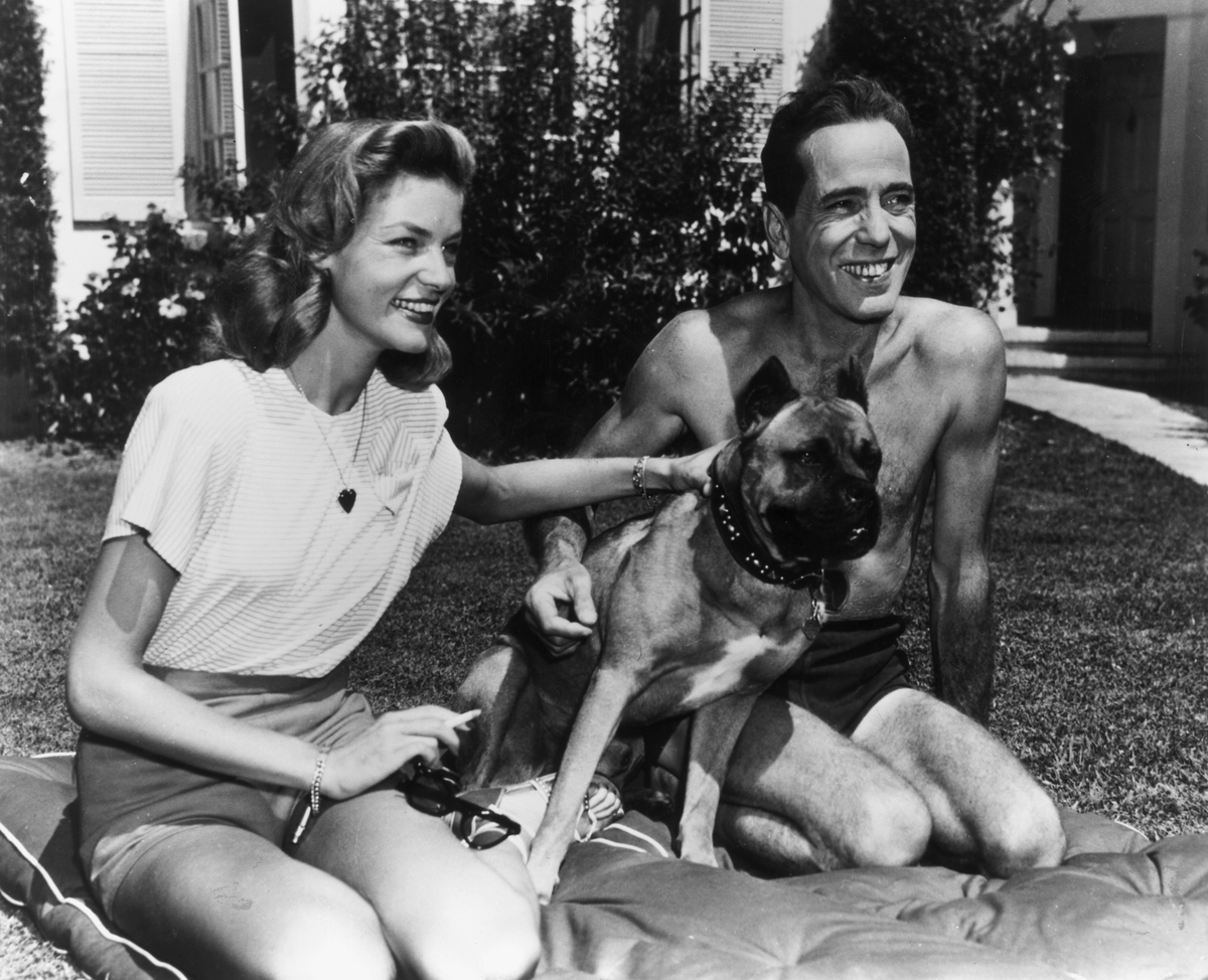 American actor Humphrey Bogart smiles as he kneels with his wife, actor Lauren Bacall, and their pet dog, on a cushion on their front lawn