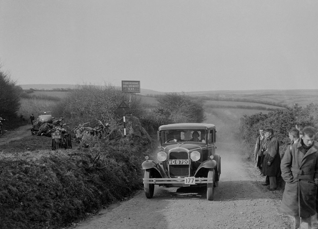 black and white photo of people driving a ford model a along a dirt road with bystanders and bikes on the side