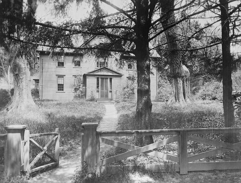 The Alcott family's wooden Orchard House is pictured in balck and white, surrounded by trees and a fence.
