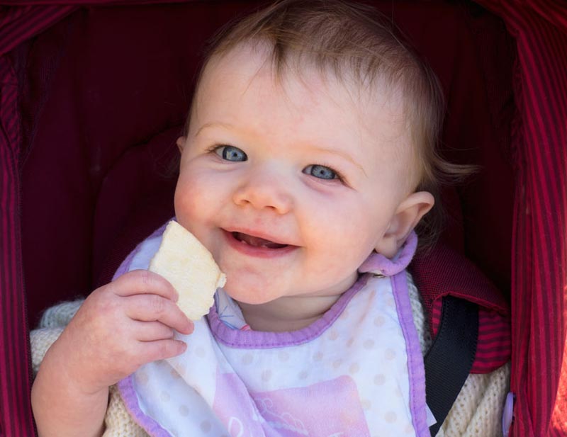 A baby smiles while holding a snack near her mouth.