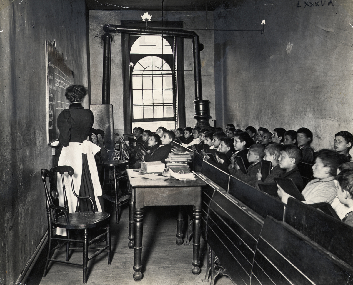A teacher demonstrates on the blackboard, as students watch attentively from crowded pews, circa 1886