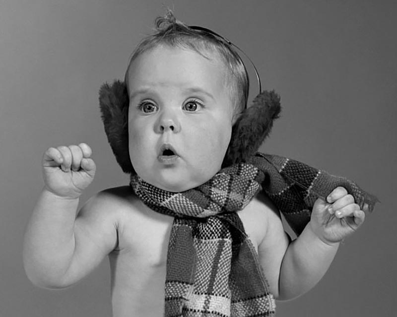 A baby wither a bear chest is dressed in ear muffs and a scarf.