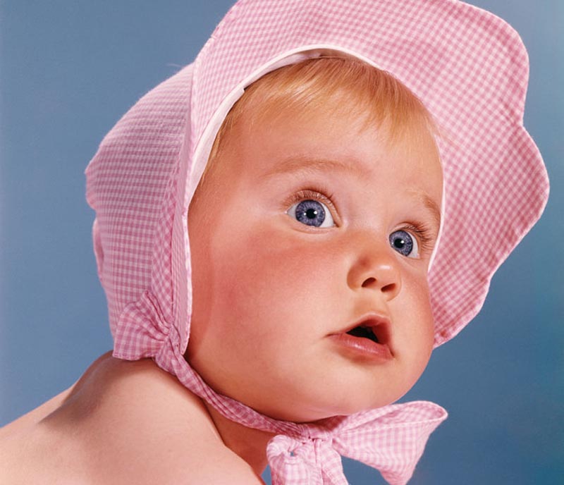 A baby in a bonnet turns to looks towards the camera.