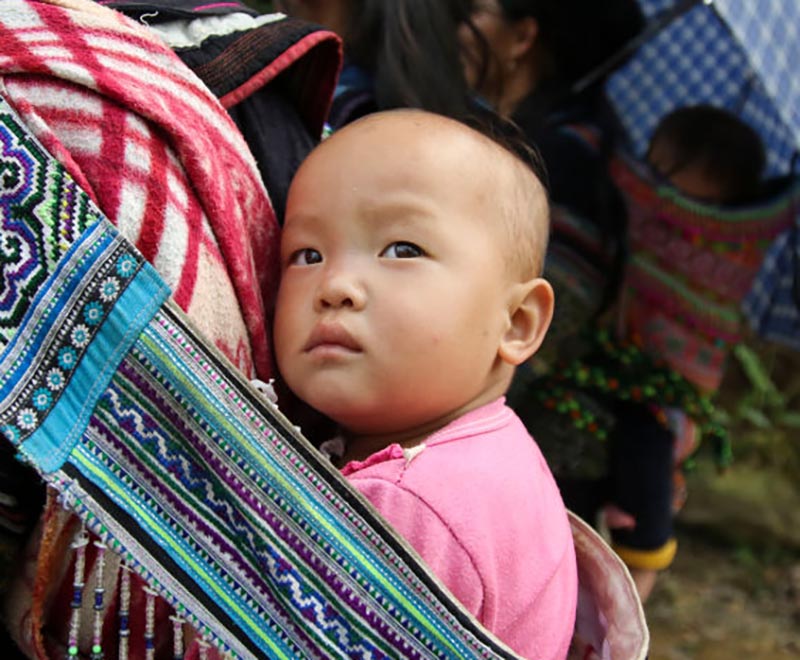 A baby is carried on someones back with a sash.