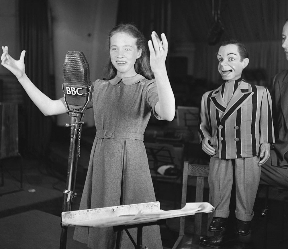 Young Julie Andrews stands in front of a microphone with her arms raised.