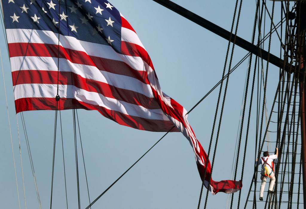 A sailor climbs the mast of the USS Constitution as an American flag waves.