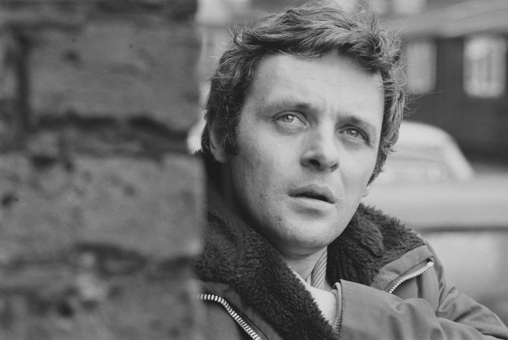 Anthony stands near a brick wall outside while reaching into his coat.