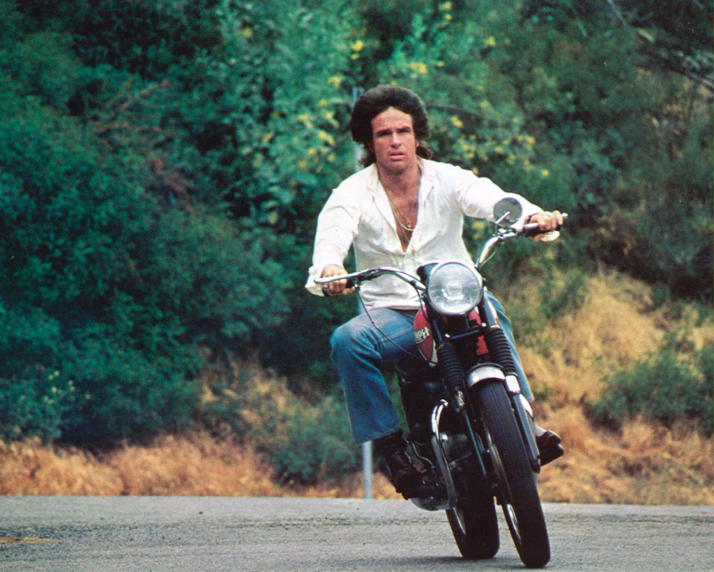 Warren Beatty drives a motorcycle near a hillside.
