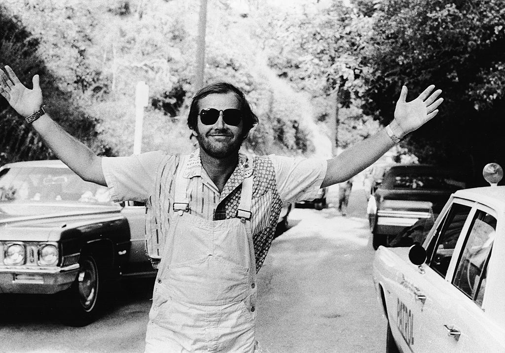 Jack Nicholson holds his arms out wide while walking through a parking lot surrounded by nature.