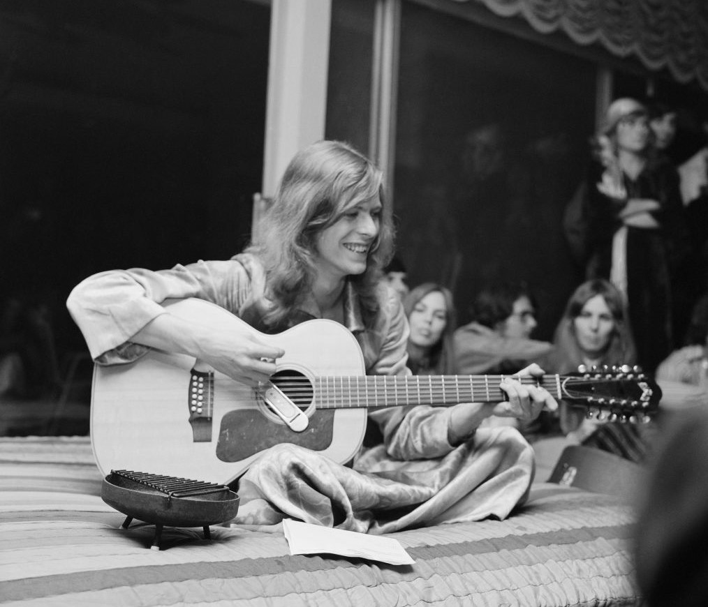 David Bowie plays guitar while sitting on a bed and surrounded by friends.