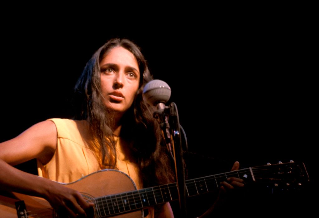 Joan Baez plays guitar onstage.