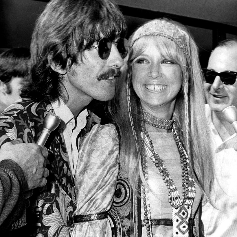 Pattie Boyd is dressed in hippie attire and smiling at George Harrison amidst reporters.