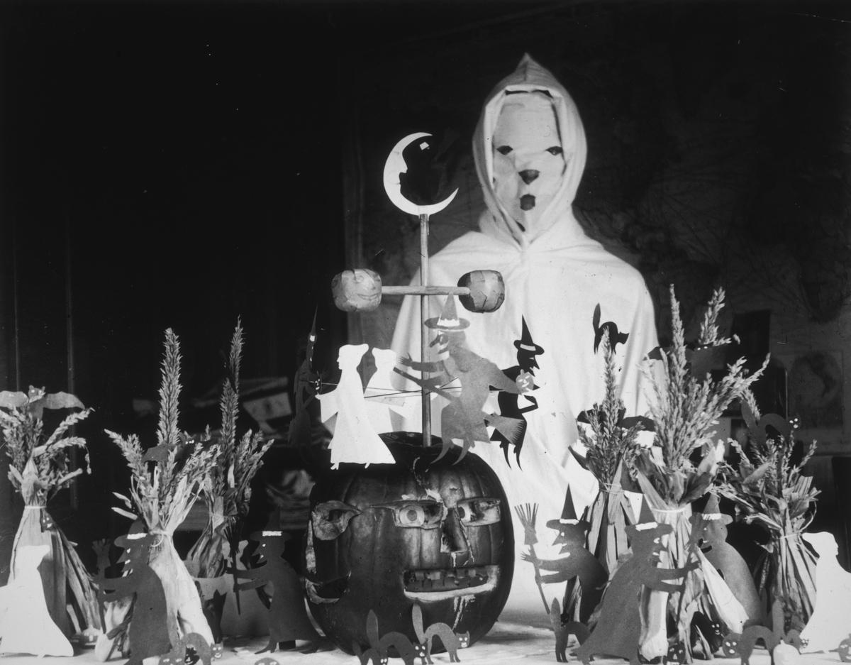 View of a person, possibly a teacher, wearing a ghost costume behind a lunch table with Halloween decorations in a rural schoolhouse. The display consists of a carved pumpkin, cutouts of witches and black cats, and haystacks.