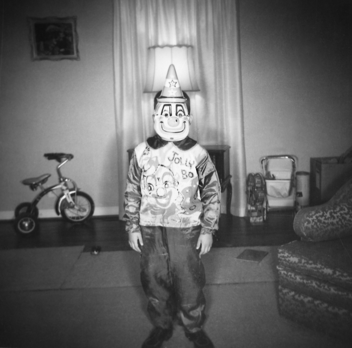 A school age boy stands in his living room for a portrait of him wearing a clown costume.