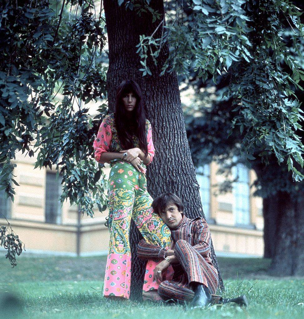 Sonny and Cher pose in pattered clothes in a park.