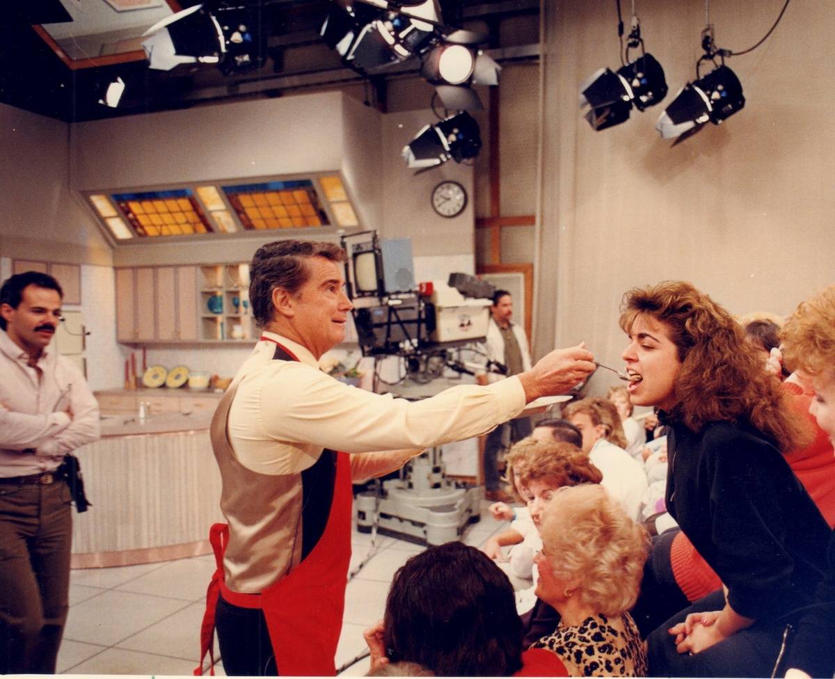 Regis Philbin feeds an audience member a sample of food from a cooking segment on The Morning Show.