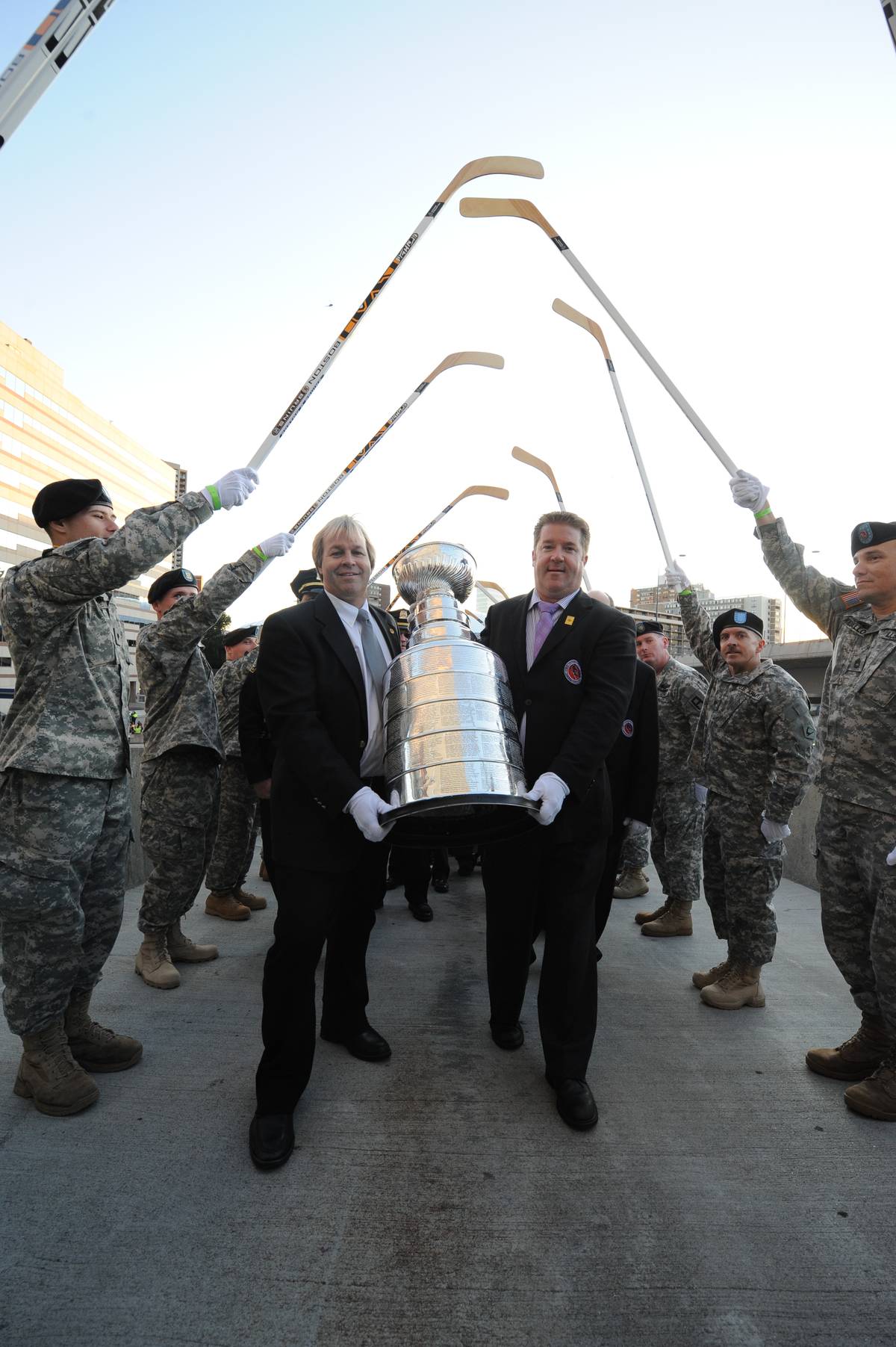 Soldiers hold up hockey sticks for Mike Bolt and Phil Pritchard to walk under while carrying the Stanley Cup.