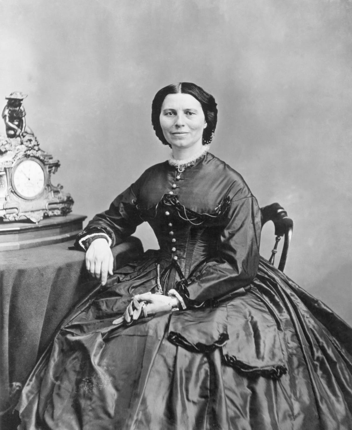 Clara Barton, under President Abraham Lincoln's charge for sending supplies to civil war soldiers, holds a pair of gloves next to a table and clock.