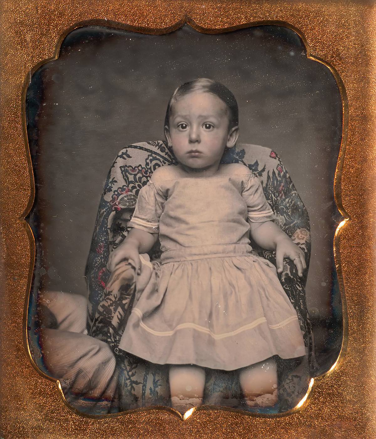 A baby is sitting on a floral chair in this 1850s photograph.