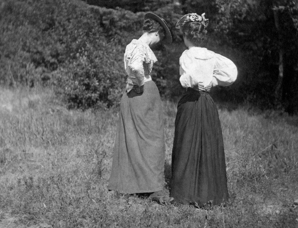 Two young women in Victorian dress talking privately with their backs to the camera