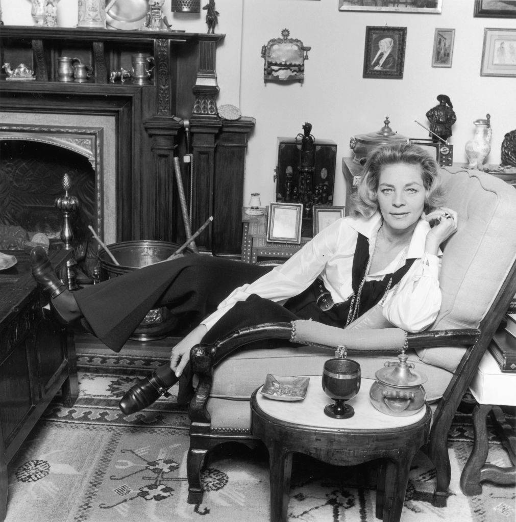 Lauren Bacall sitting in an armchair with one leg propped on a table, at her home