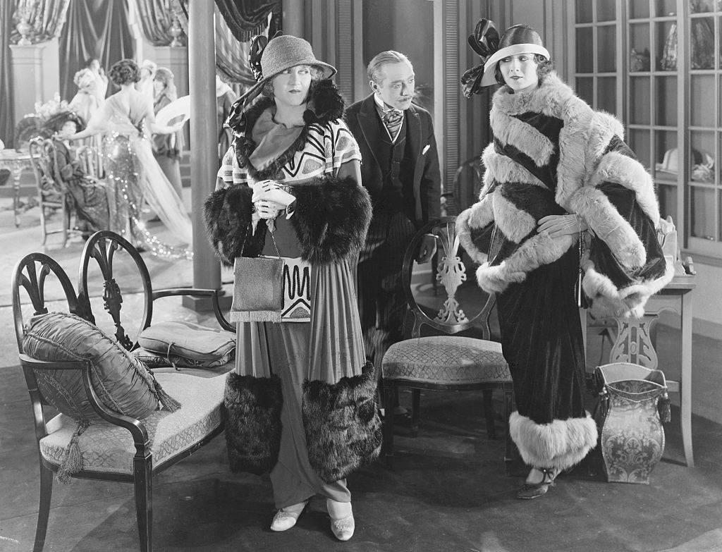 Two women, visiting a dress salon, donning styles of the late 1900s, including broad-brimmed, ribboned hats, fur trimmed cloaks over ankle-length, slim-lined, hobble-skirt dresses, clutching purses with gloved hands, and wearing high-heeled shoes