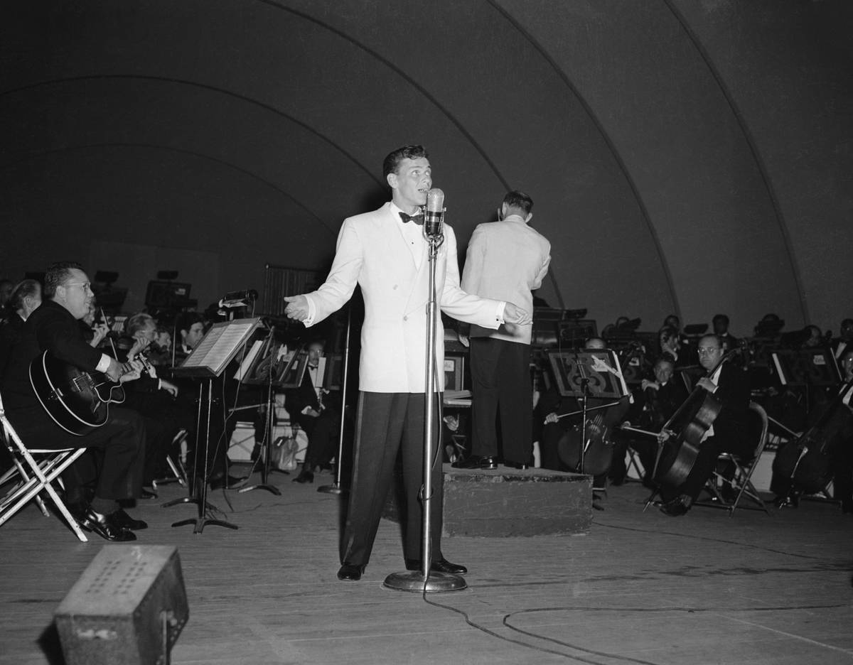 Frank Sinatra performs at the Hollywood Bowl. Here he stands full length crooning into a microphone, wearing a white jacket and black pants.