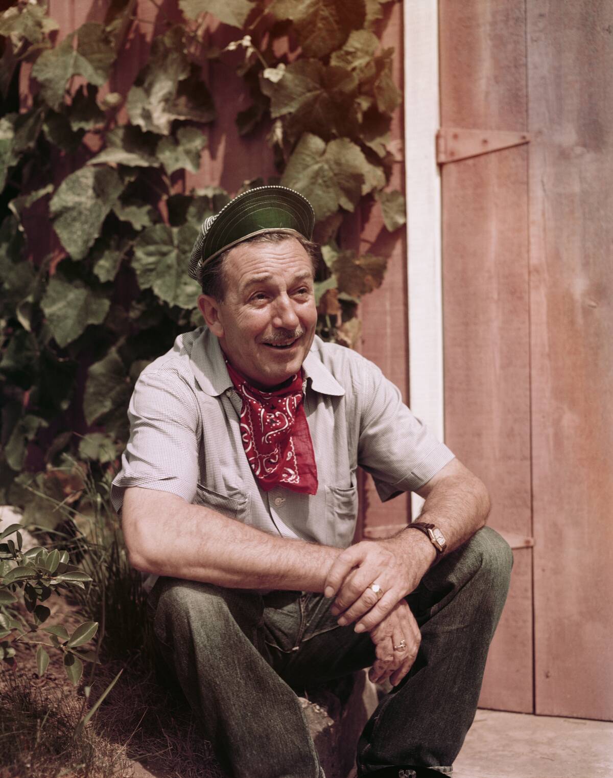 Portrait of American movie producer, artist, and animator Walt Disney (1901 - 1966) as he sits outdoors beside a wooden door, California,
the early to mid-1950s. He wears a train engineer's cap and a red
neckerchief.
