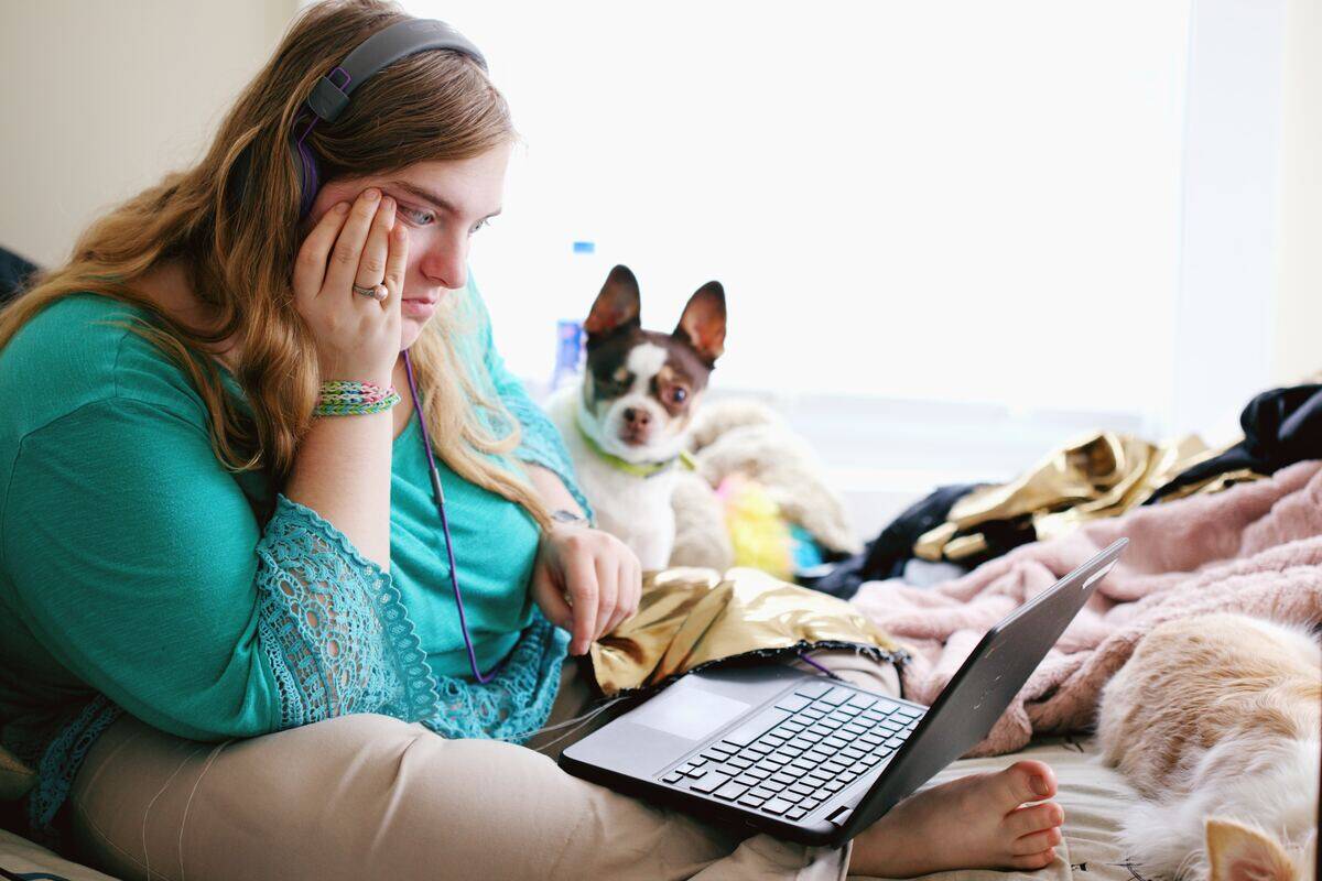 woman in headphones looks frustrated while using laptop on bed as dog looks on