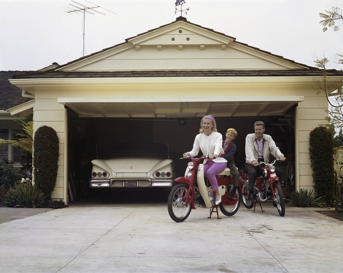 Family sitting on Honda 50 mopeds in front of garage, smiling