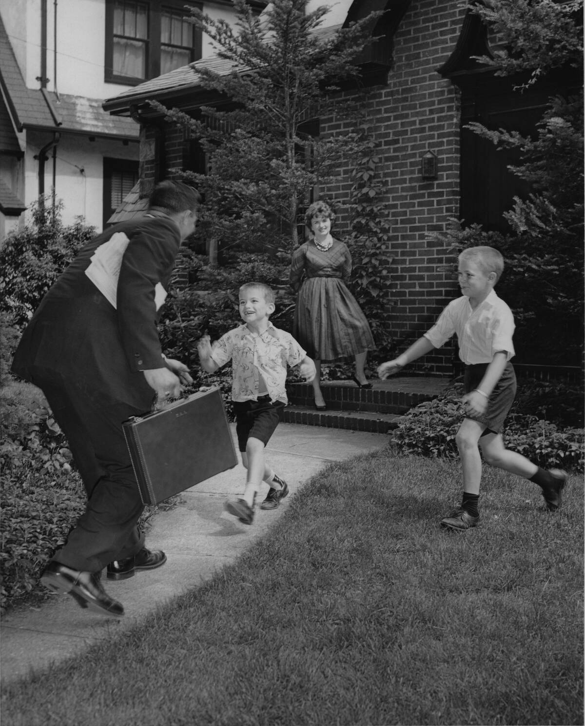 Two Boys Running To Meet Dad Coming Home From Work While Mom Looks On