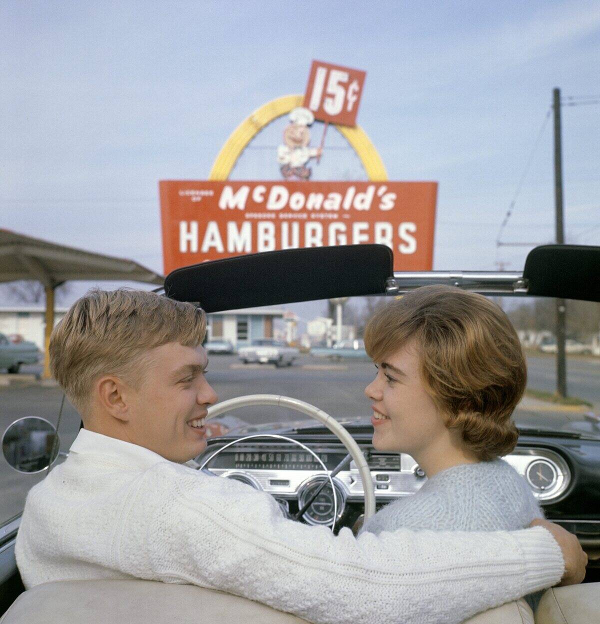 Fast food in the 1950s. A young couple in their convertible car with a McDonald's sign in the backgrou...