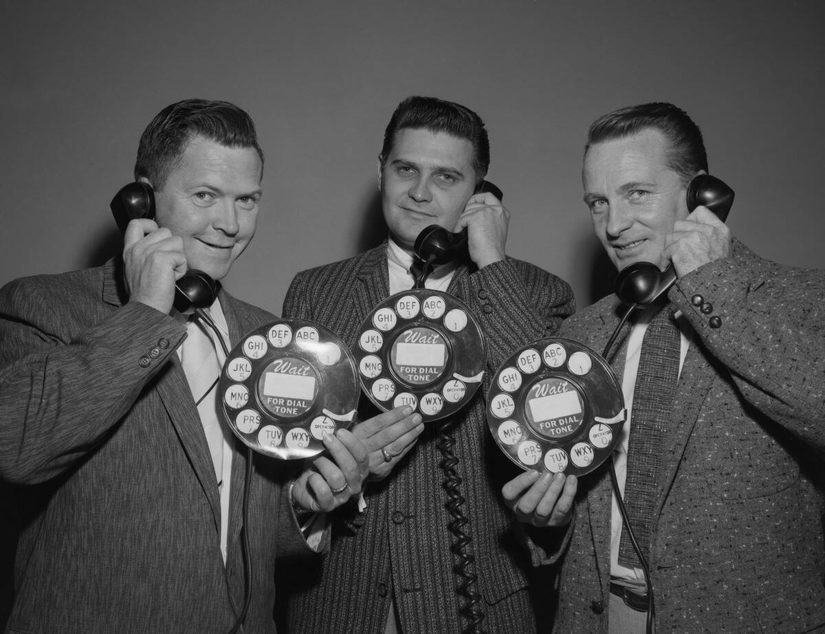Three men happily hold enlarged rotary telephone dials while on the phone, ca. 1955.