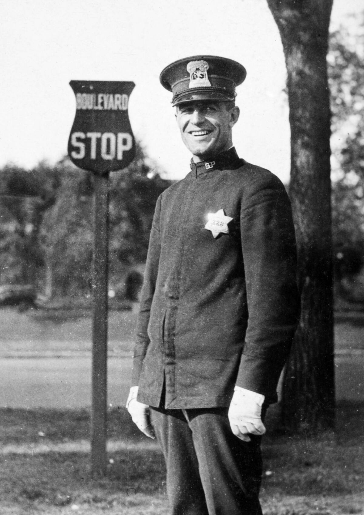 Policeman smiles by a bus stop sign, ca. 1925