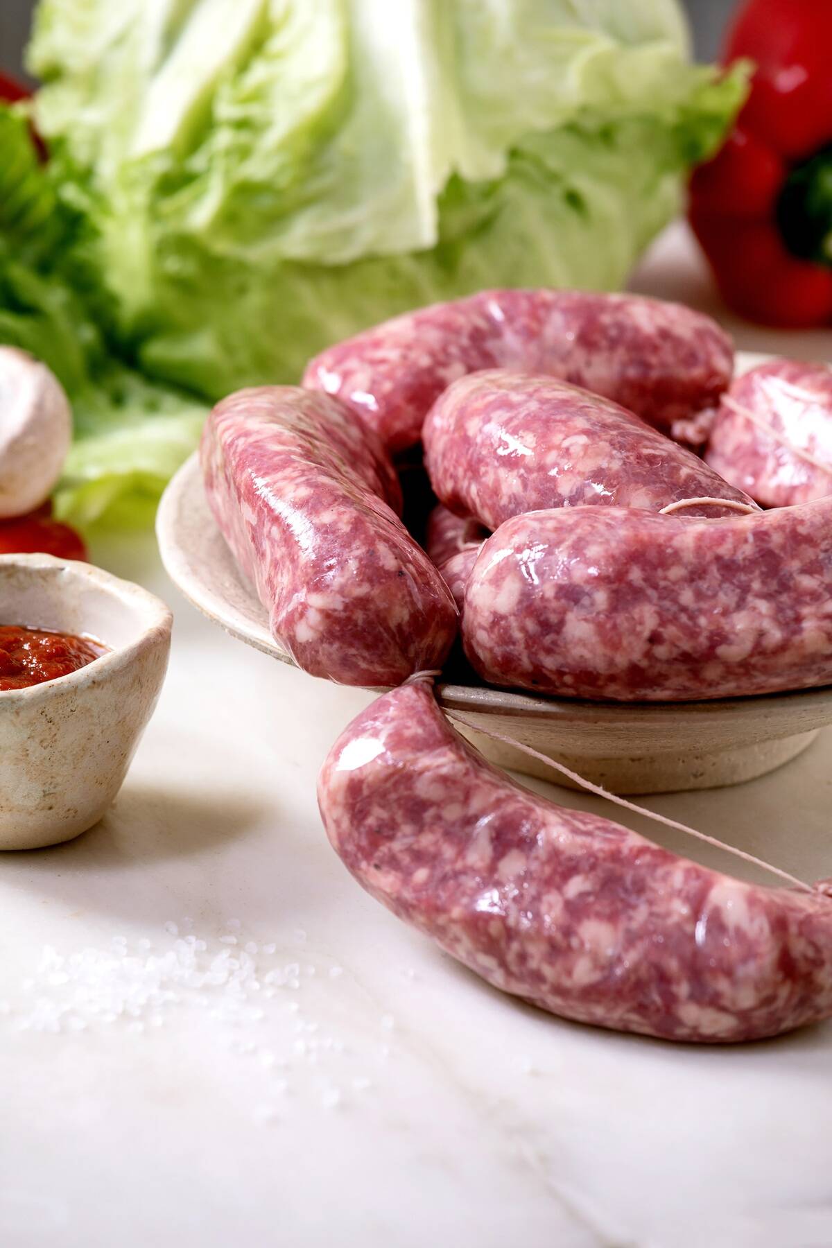 Raw uncooked italian sausages salsiccia in plate on white marble table. Green salad. vegetables and tomato sauce around.