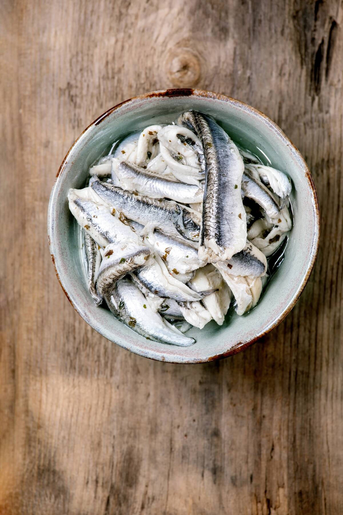 Bowl of pickled anchovies or sardines fillet in oil with herbs for tapas or antipasti over old wooden background. Flat lay, copy space
