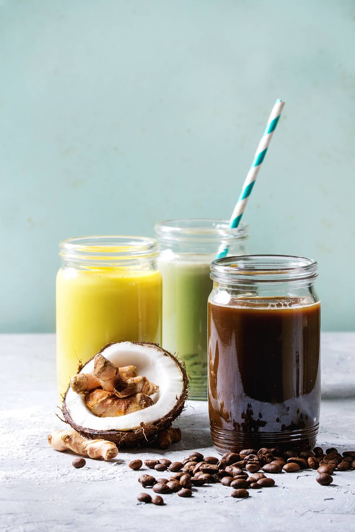 Variety of iced colorful latte drinks Iced coffee, turmeric and matcha latte cocktails in glass jars with ingredients above over grey green texture background Copy space