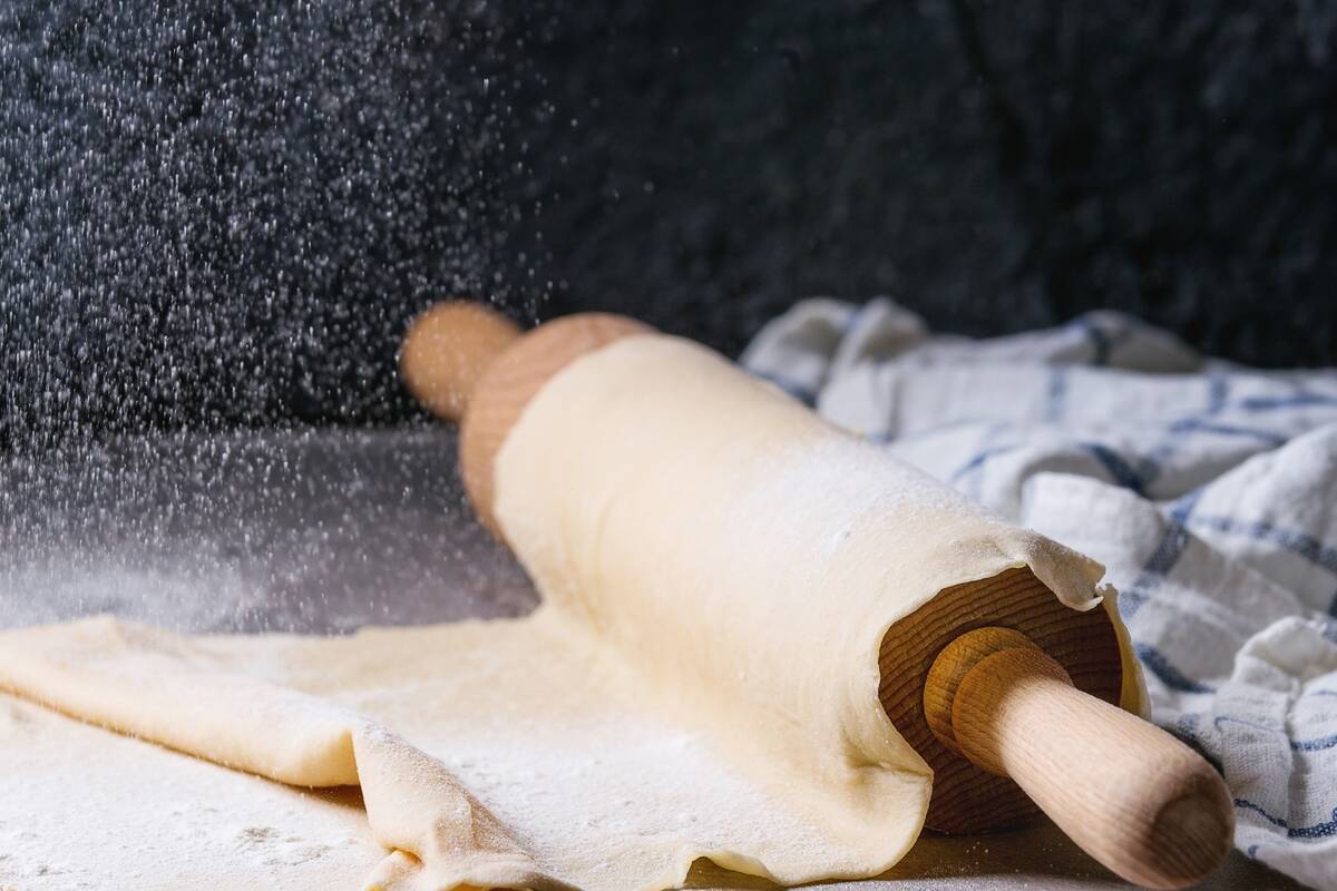 Fresh homemade rolled dough for pasta tagliatelle on wood rolling pin with sieve sprinkling flour and kitchen towel over dark gray table background.