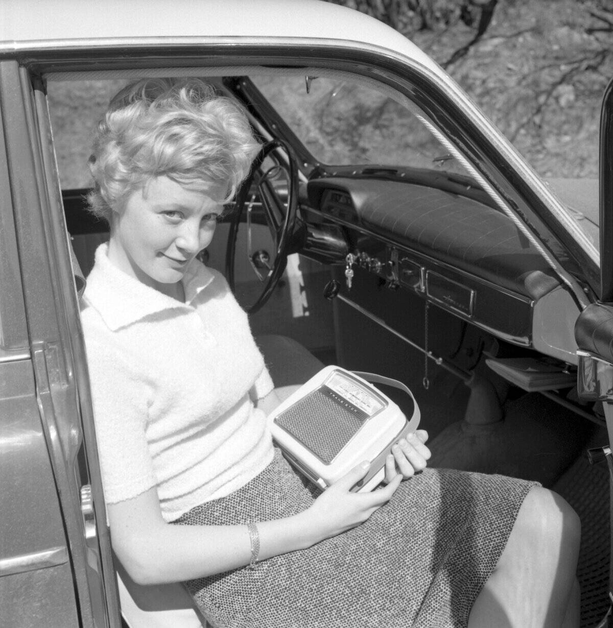 In the 1960s. A young woman in a car listening to a portable radio. Battery operated transistor radio ...