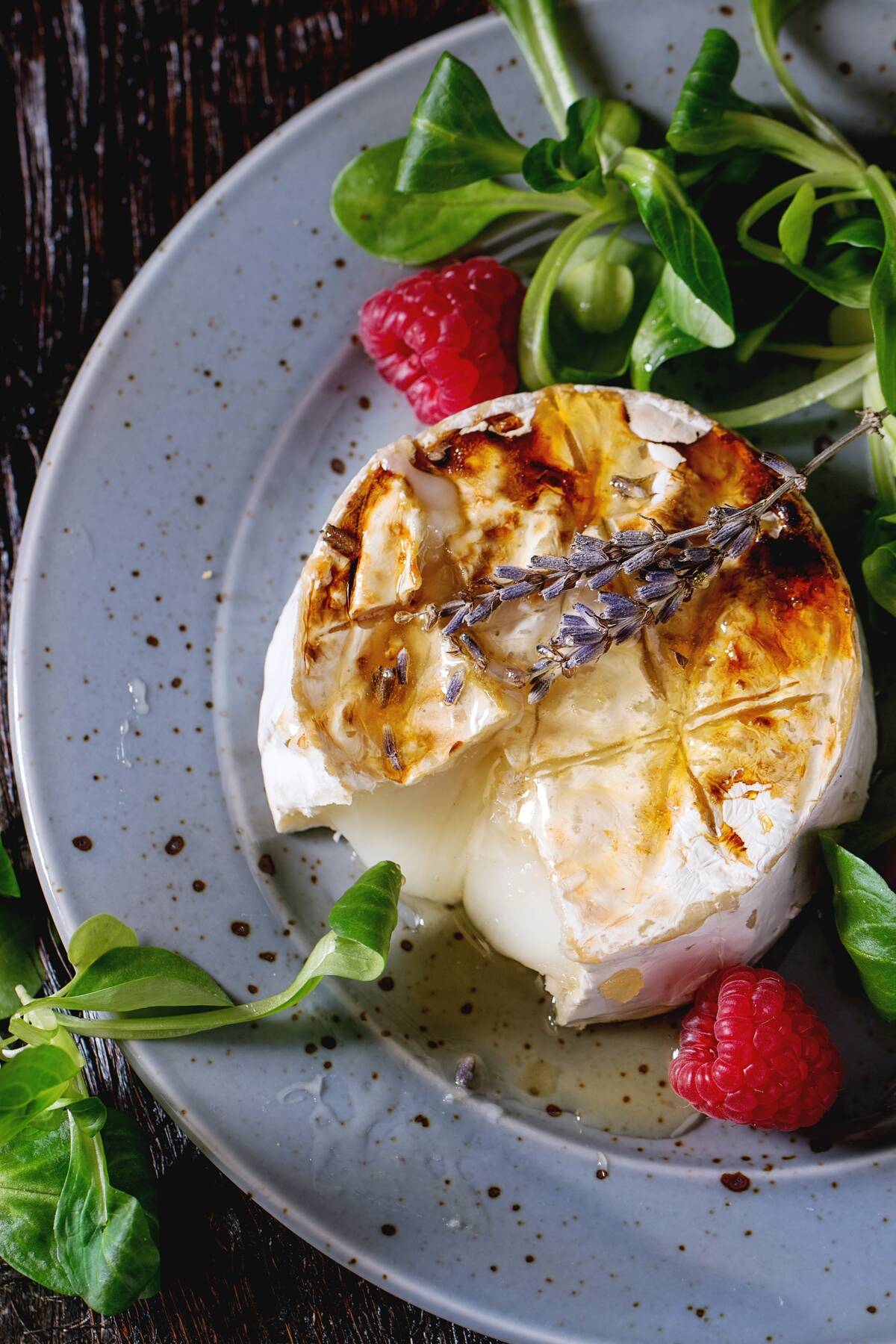 Melting sliced grilled goat cheese on blue plate, served with liquid honey, lavender, raspberries, wholegrain toast and green salad. Over old wooden table. Dark rustic style. Top view